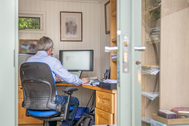 Man working at a computer inside a bright, white-panelled garden office, viewed through the open doors.